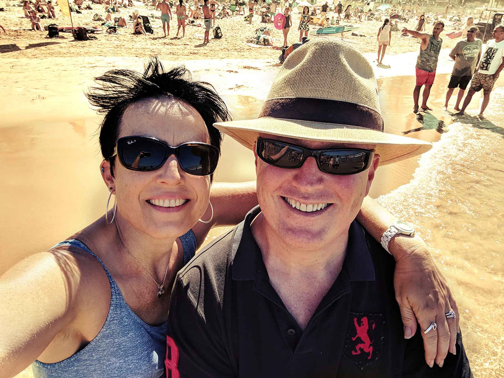 A smiling couple in sunglasses taking a selfie on a busy sunlit beach