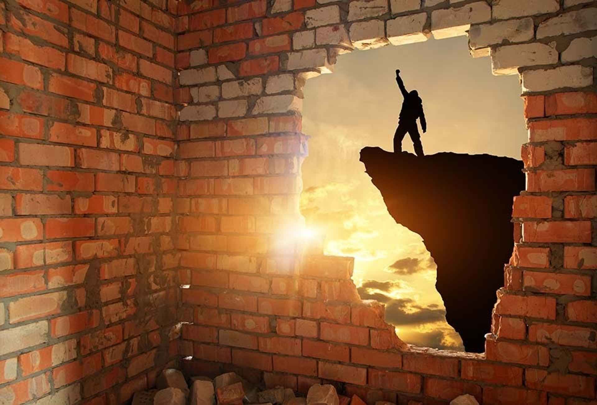 A silhouetted figure raising a fist in triumph on a clifftop at sunset, seen through a hole in a brick wall