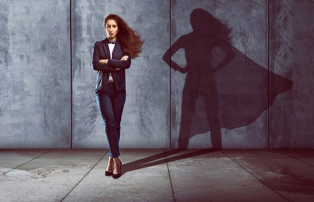 A businesswoman standing confidently against a concrete wall, her shadow cast as a caped superhero