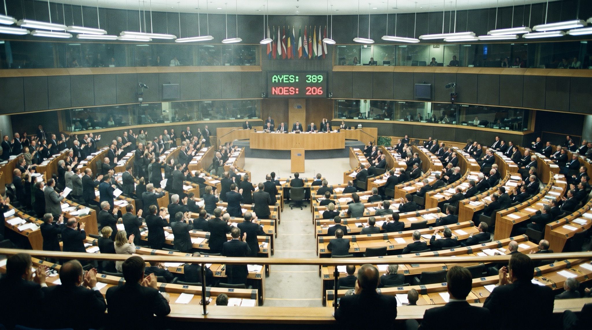 A modern parliamentary debating chamber seen from a high rear balcony, its semicircular tiers of pale wood desks split down the middle mid-vote