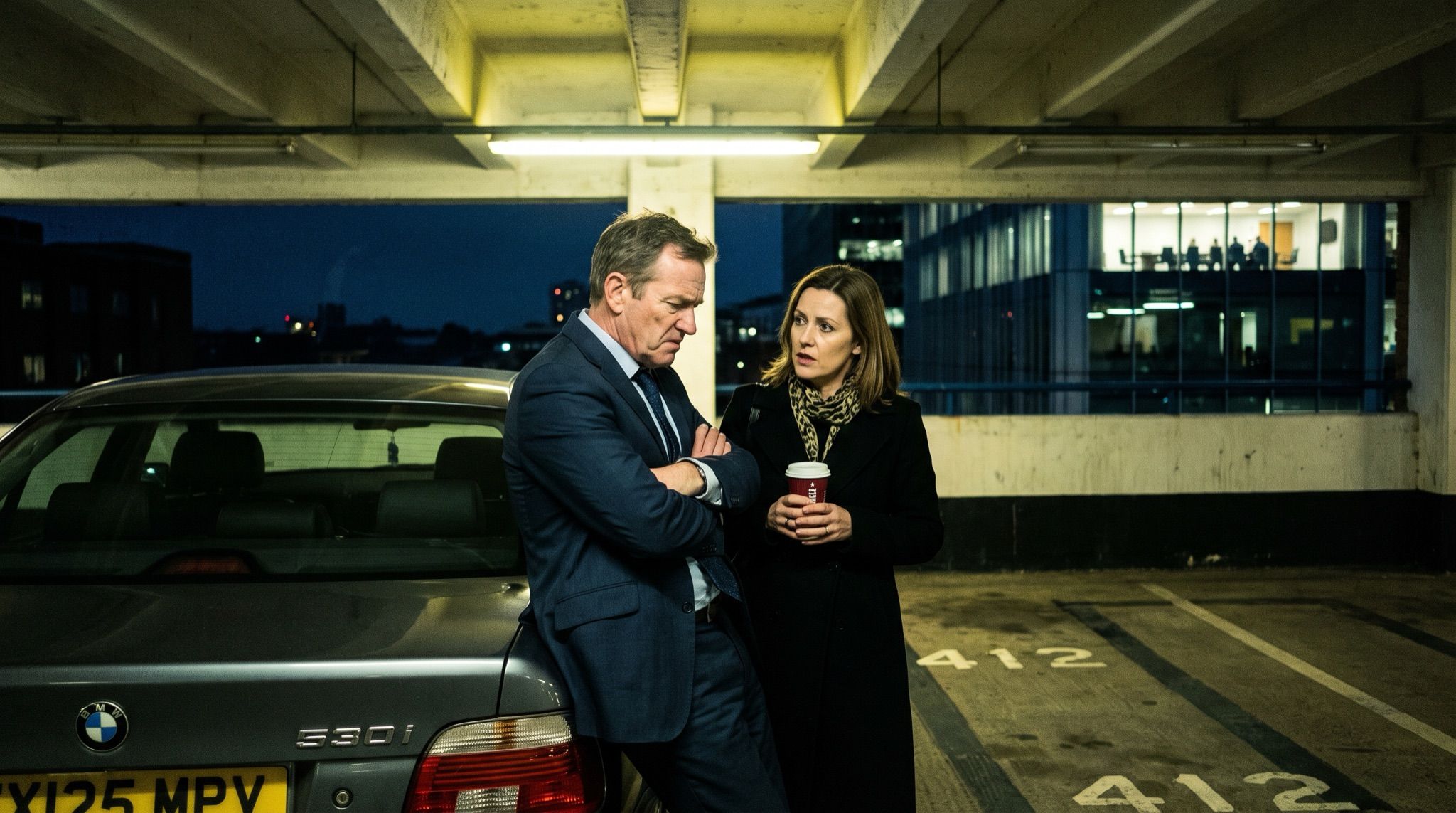 Two colleagues in business dress standing close together at night in a bare concrete corporate car park, under hard overlapping strip light